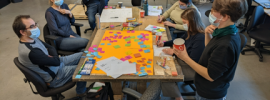 A group of people are sitting around a table covered in post-its and a large sheet of orange paper. One person on the right is standing.