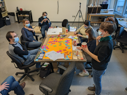 A group of people are sitting around a table covered in post-its and a large sheet of orange paper. One person on the right is standing.